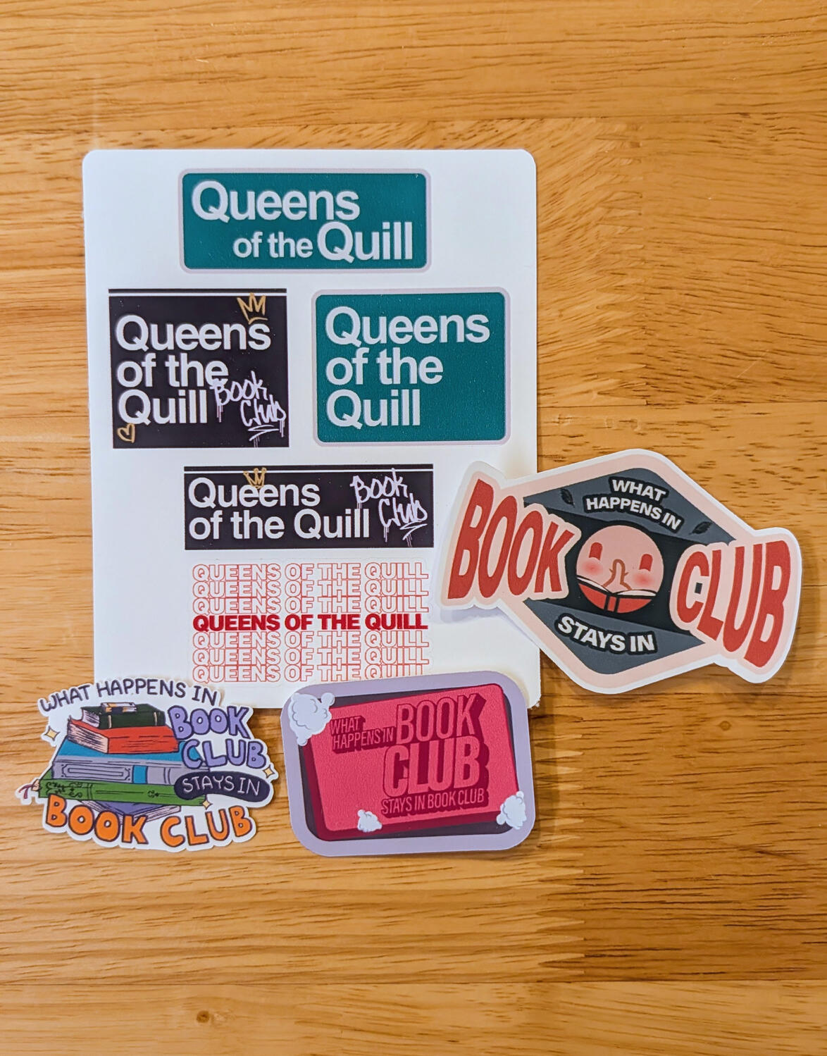 A sticker sheet with variations of "Queens of the Quill" done in NYC style signage. Around the sheet has various sticker designs that read "What happens in book club stays in book club."